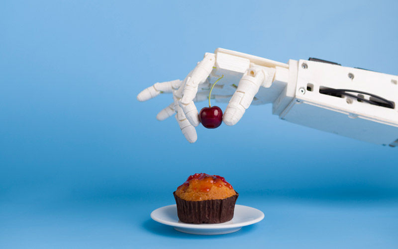 Ein Roboterarm legt eine Kirsche auf einen Muffin